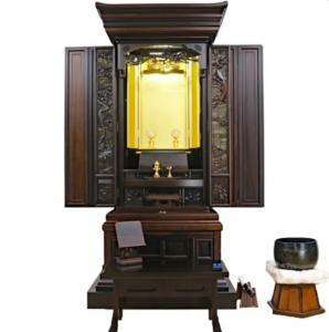 Traditional dark wood butsudan Buddhist altar with doors open to a gold inner shrine, offerings on the altar, and a prayer book on a nearby table with a bowl on the right.