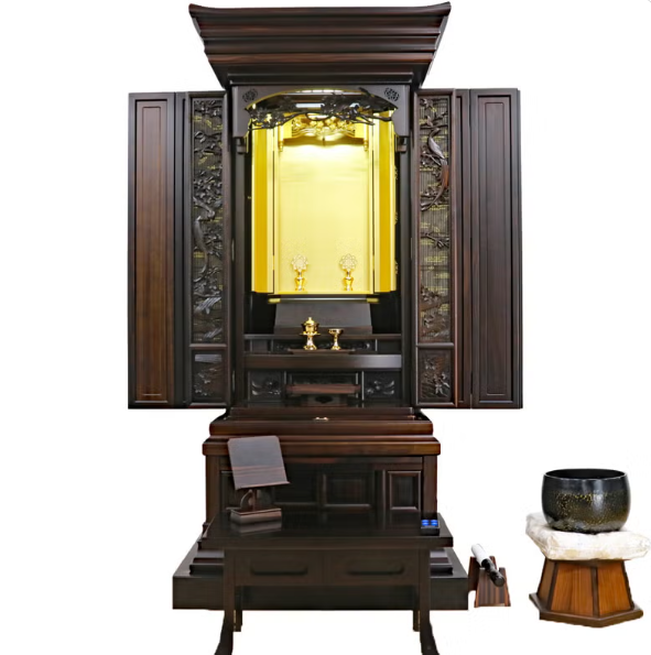 Traditional dark wood butsudan Buddhist altar with doors open to a gold inner shrine, offerings on the altar, and a prayer book on a nearby table with a bowl on the right.