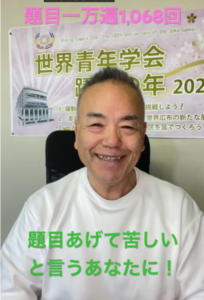 Smiling man in a white shirt sits in front of a banner with Japanese text and floral graphics; green text printed across his shirt reads '題目あげて苦しいと言うあなたに！'