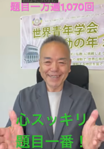 Smiling man in a gray blazer seated in front of a banner with Japanese text; pink caption at top, green caption at bottom conveys a positive message.