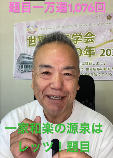 Smiling man in a white shirt speaks to the camera, with a poster behind him and pink Japanese caption at the top and green caption at the bottom