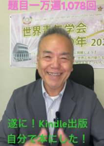 満面の笑みを浮かべる男性が椅子に座り、背後の横断幕と緑の文字で『Kindle出版 自分で本にした！』と宣伝している。戻る背景にはイベントの旗やロゴも見える。
