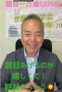 Smiling Japanese man in a gray blazer seated, with a banner containing Japanese text behind him.