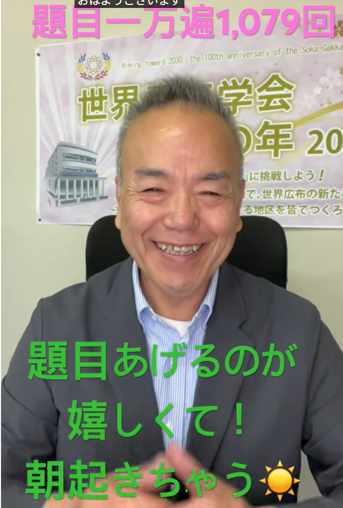 Smiling Japanese man in a gray blazer seated, with a banner containing Japanese text behind him.