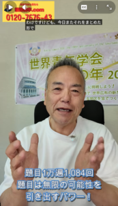 Smiling man in a white shirt speaks to the camera, hands raised; a banner and poster are visible behind him with blue subtitle text at the bottom.