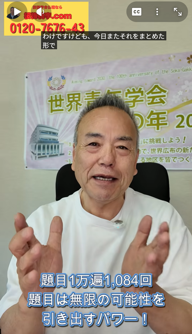 Smiling man in a white shirt speaks to the camera, hands raised; a banner and poster are visible behind him with blue subtitle text at the bottom.