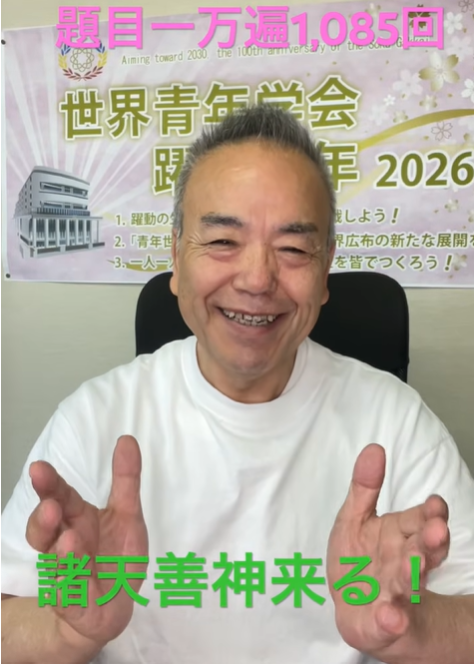 Smiling man in a white shirt raises his hands in front of a banner for a 2026 event, with pink text across the top and green text at the bottom.