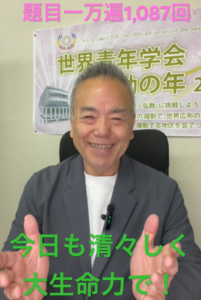 笑顔の男性が椅子に座り前方を見て話している。背景にはピンクの文字『題目一万遍1,087回』と緑の文字『今日も清々しく 大生命力で！』の字幕がある。