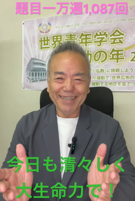 笑顔の男性が椅子に座り前方を見て話している。背景にはピンクの文字『題目一万遍1,087回』と緑の文字『今日も清々しく 大生命力で！』の字幕がある。