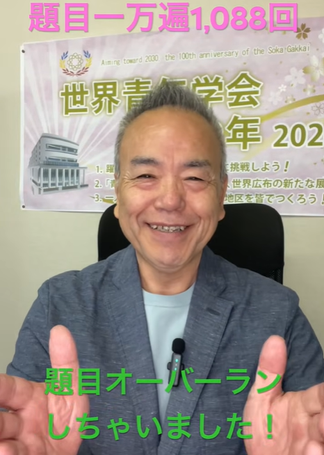 Smiling man in a blue blazer shows thumbs up while seated in front of a banner with Japanese text and a logo in a conference setting.