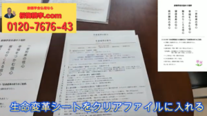 Desk with printed documents on a dark table and a yellow top-left banner; blue caption at bottom instructs to put the life-change sheet in a clear file.