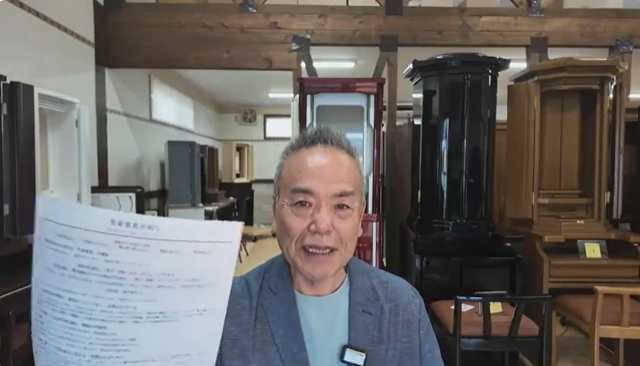 Smiling man in a blue blazer holds a document toward the camera in a showroom filled with wooden furniture pieces behind him.