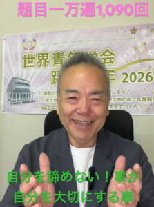Smiling man in a blazer facing the camera with a banner behind him and green Japanese captions overlaid.