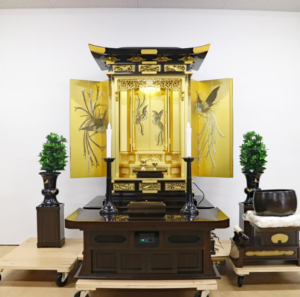 Open gold Buddhist altar shrine (butsudan) on a dark wooden table, with two tall candle holders and greenery on each side.