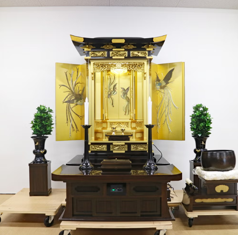 Open gold Buddhist altar shrine (butsudan) on a dark wooden table, with two tall candle holders and greenery on each side.