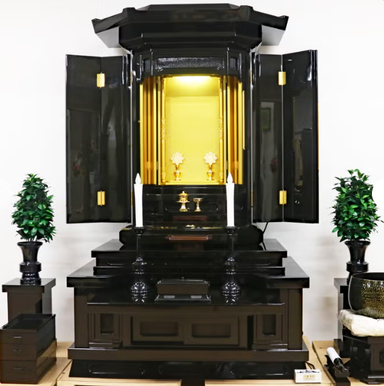 Black lacquered home altar with doors open to reveal a gold inner shrine, candles, and ritual items on the steps. Flanked by potted plants.