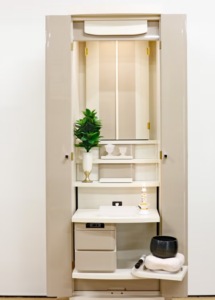 Cream home altar cabinet with plant, candle, bowls, and small workspace inside.