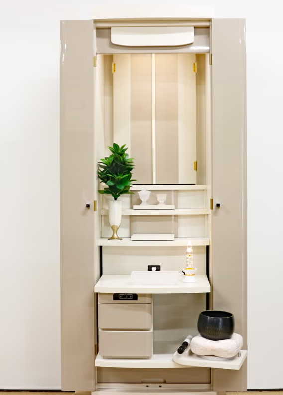 Cream home altar cabinet with plant, candle, bowls, and small workspace inside.