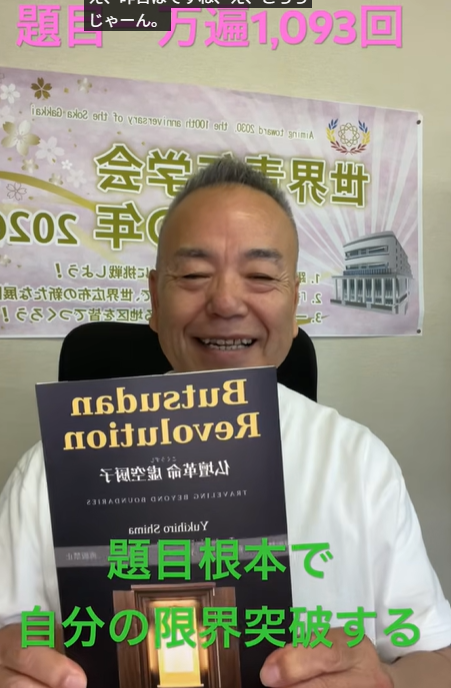 Smiling man in a white shirt holds a dark blue book with gold lettering, seated indoors with a purple Japanese banner in the background.