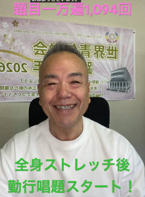 Smiling man in a white shirt seated in front of a banner with Japanese text promoting a full-body stretch and chant session.