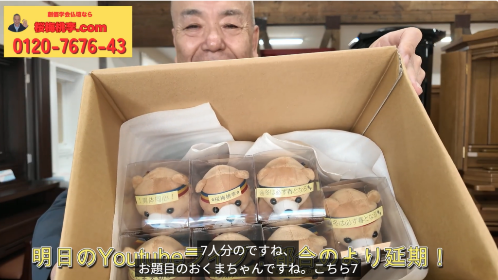 A person smiles while holding an open box containing six small teddy bear plushes in clear plastic cases; a yellow ad banner with Japanese text and a phone number sits in the top-left corner.