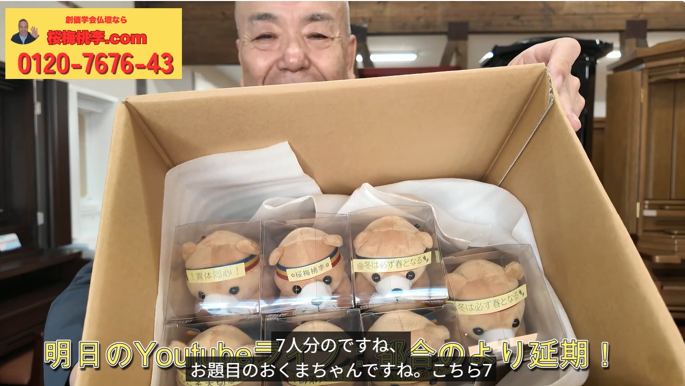 A person smiles while holding an open box containing six small teddy bear plushes in clear plastic cases; a yellow ad banner with Japanese text and a phone number sits in the top-left corner.