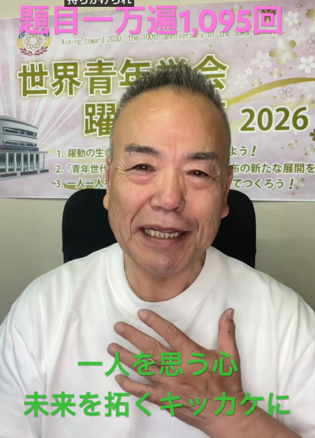 Smiling man in a white shirt speaks to the camera with a Japanese event banner behind him mentioning 2026.