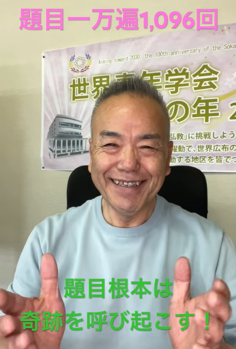 Smiling man in a light blue shirt sits in front of a banner with Japanese text; pink caption at top and green caption near bottom of the image convey the message.