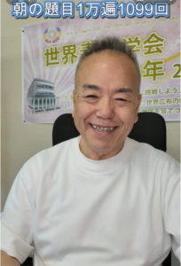 Smiling Asian man in a white shirt sits in front of a poster with Japanese text and a building illustration in the background.