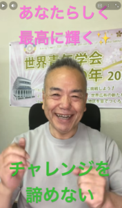 Smiling man in a bright room with a banner behind him; pink text reads 'あなたらしく 最高に輝く' and green text 'チャレンジを諦めない'.
