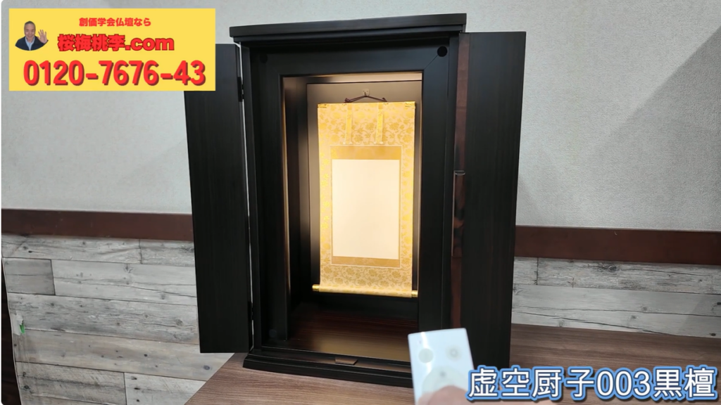 Black wooden altar (butsudan) with an ornate gold hanging scroll inside, on a wooden table; a TV remote is visible in the foreground.