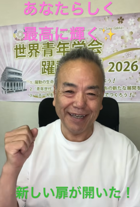 Smiling man in a white shirt with his fist raised, seated in front of a colorful banner advertising a 2026 event.