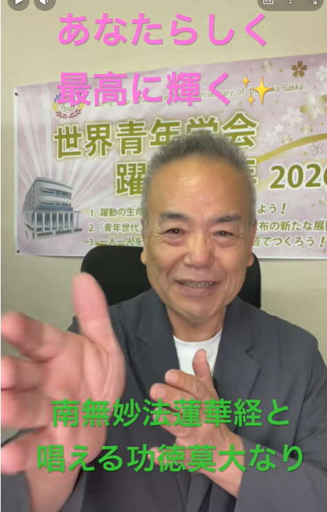 Smiling man in a blazer speaks to the camera, with a poster/banner in the background and pink and green Japanese captions overlaid.