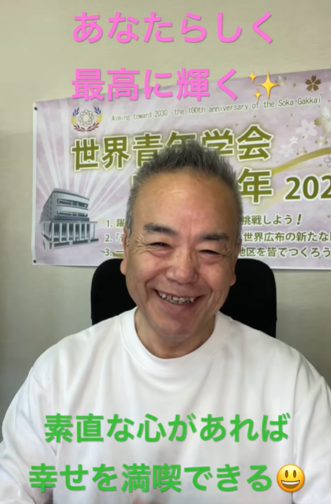 Smiling man in a white shirt sits indoors in front of a purple event banner; pink caption at top and green caption at bottom convey a positive message in Japanese.