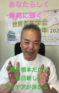 Smiling man in a white shirt sits in front of a banner with pink and green Japanese captions overlaid: 'あなたらしく 最高に輝く' and '題目根本だから 毎日新しい アイデアが浮かぶ', hands raised as if explaining.