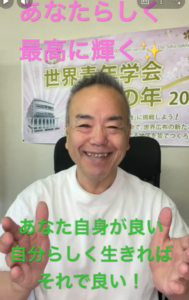 Smiling man in a white shirt sits in front of a banner with Japanese text; bright pink caption appears at the top and green caption at the bottom.