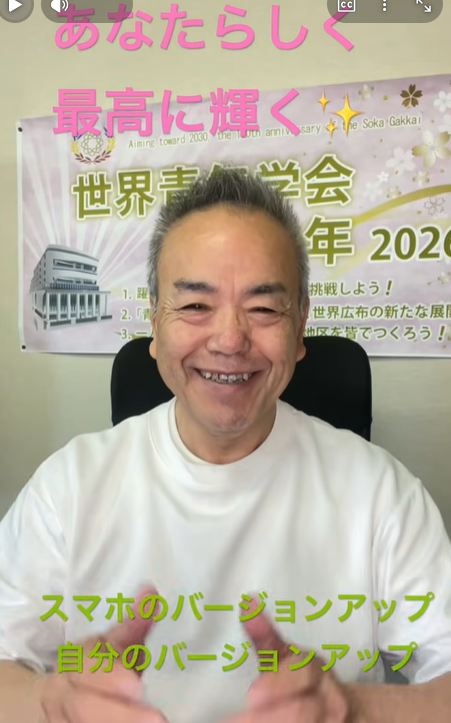 Smiling man on a video call with a Japanese event poster behind him; green caption at bottom reads 'スマホのバージョンアップ 自分のバージョンアップ'.