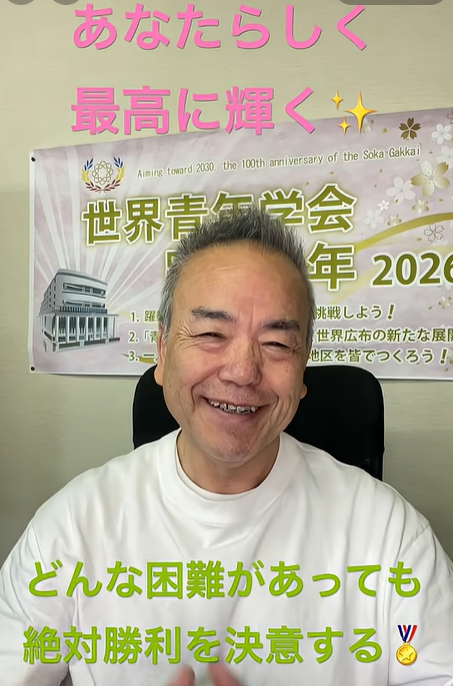 Smiling man in a white shirt sits in front of a banner with Japanese text, pink slogans above and green text at the bottom about staying true and overcoming challenges.