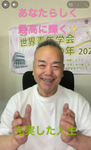 Smiling man in a white shirt sits in front of a poster with Japanese text; pink heading above and green text below.