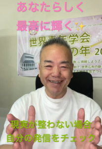 Smiling man in a white shirt facing the camera, hands raised, with a banner behind him and pink text above: 'あなたらしく 最高に輝く' and green text below: '現実が整わない場合 自分の発信をチェック'.