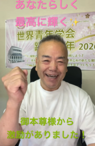 Cheerful man in a white T-shirt pumps his fist at the camera, with pink and green Japanese text banners behind him.