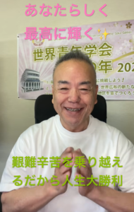 Smiling man in a white shirt sits in front of a Japanese event banner with pink and green text overhead and below him.