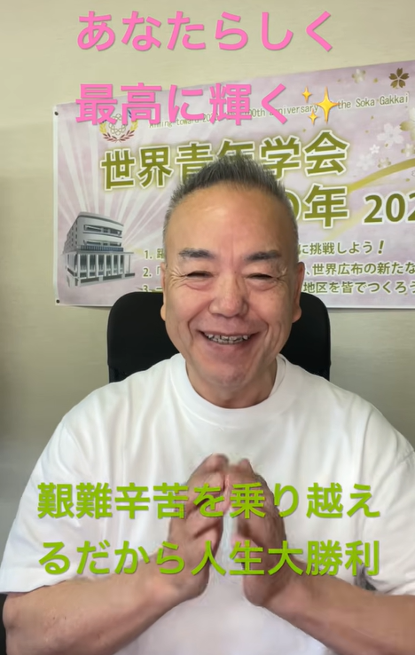 Smiling man in a white shirt sits in front of a Japanese event banner with pink and green text overhead and below him.