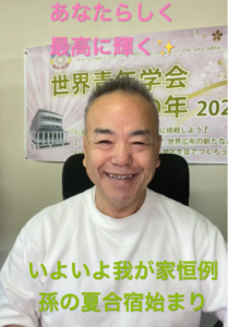 Smiling Asian man in a white shirt sits in front of a poster with pink text at the top and green text at the bottom in Japanese.