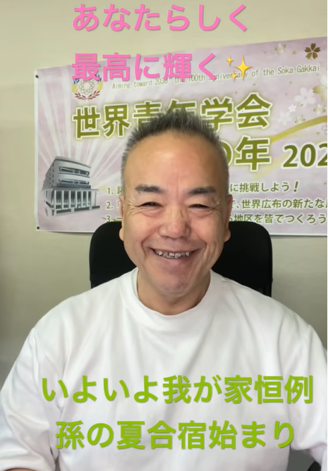 Smiling Asian man in a white shirt sits in front of a poster with pink text at the top and green text at the bottom in Japanese.