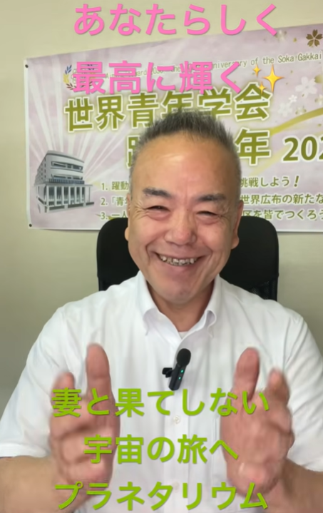 Smiling man in a white shirt with a clip-on mic sits in front of a Japanese event banner with pink and green caption text about space travel and a planetary observatory.