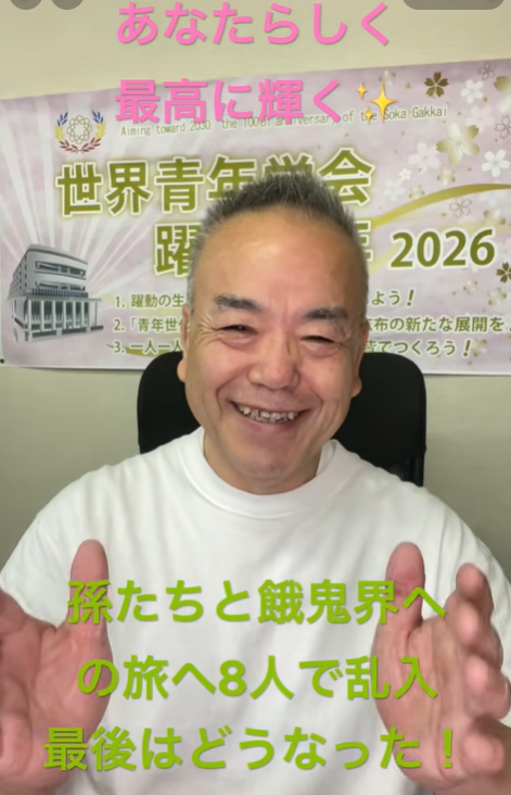 Smiling man in a white shirt sits in front of a Japanese poster, gesturing with open hands toward the camera.