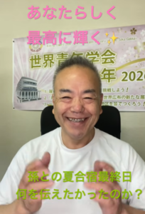 Smiling man in a white shirt speaks in front of a banner with Japanese text; pink caption above and green caption below.