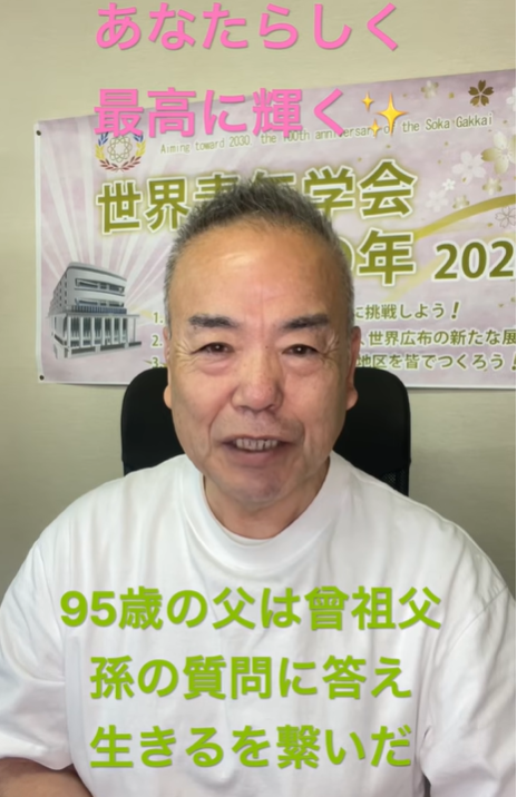 Smiling Japanese man in a white shirt speaks to the camera, seated in front of a poster with pink headline text above and green caption text below.