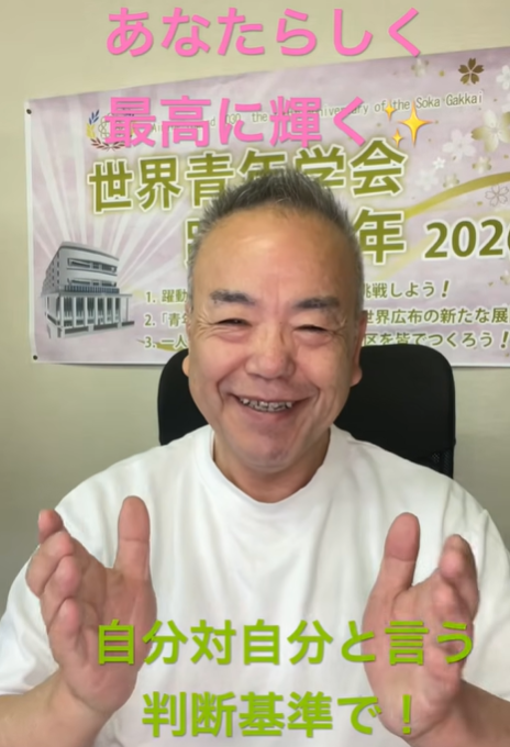 Smiling man in a white T-shirt facing the camera, hands raised, sitting in front of a banner with Japanese text and a building illustration in the background.